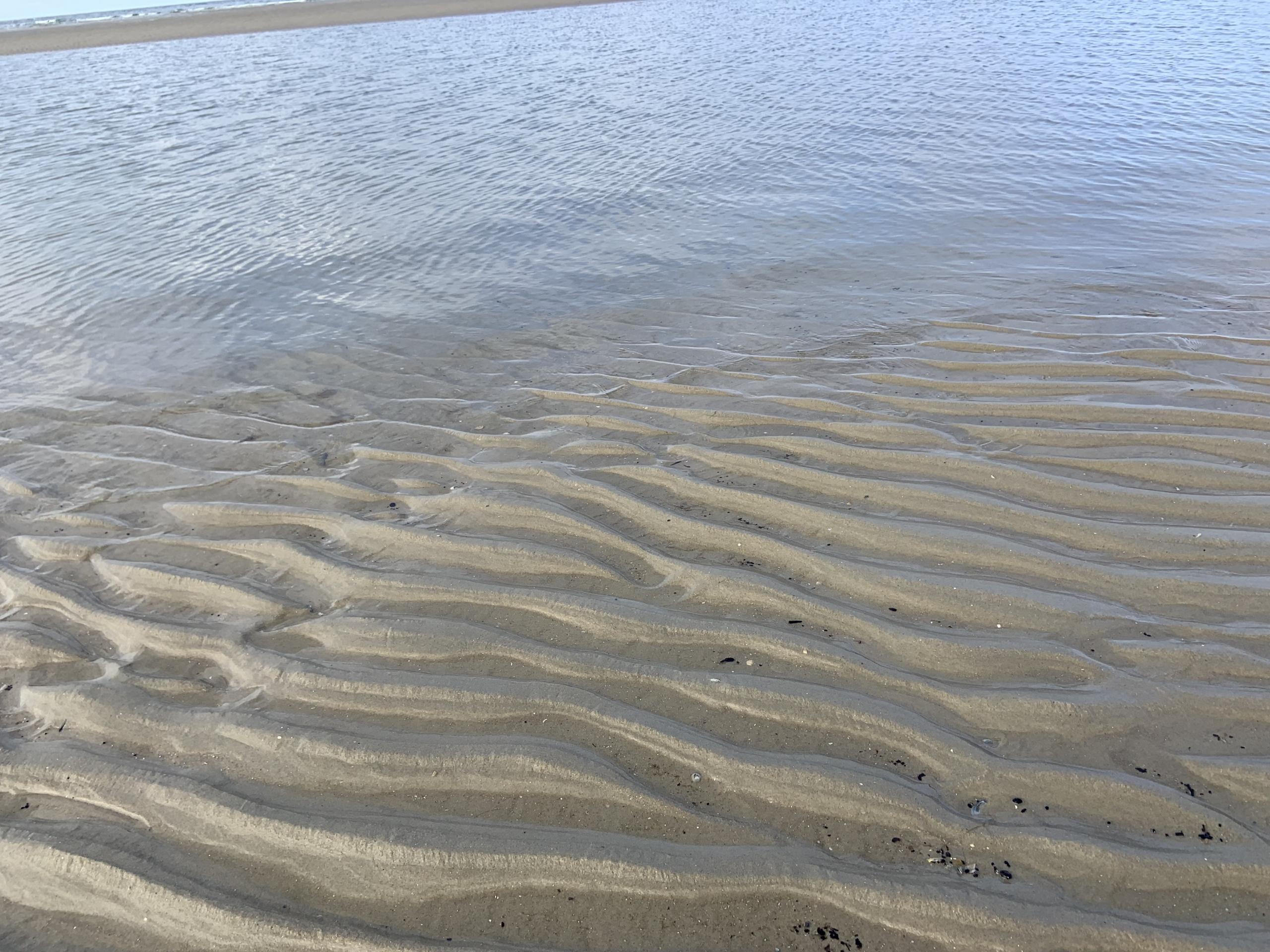 Flaches Wasser am Strand mit wellenförmigen Sandstrukturen unter der Wasseroberfläche.